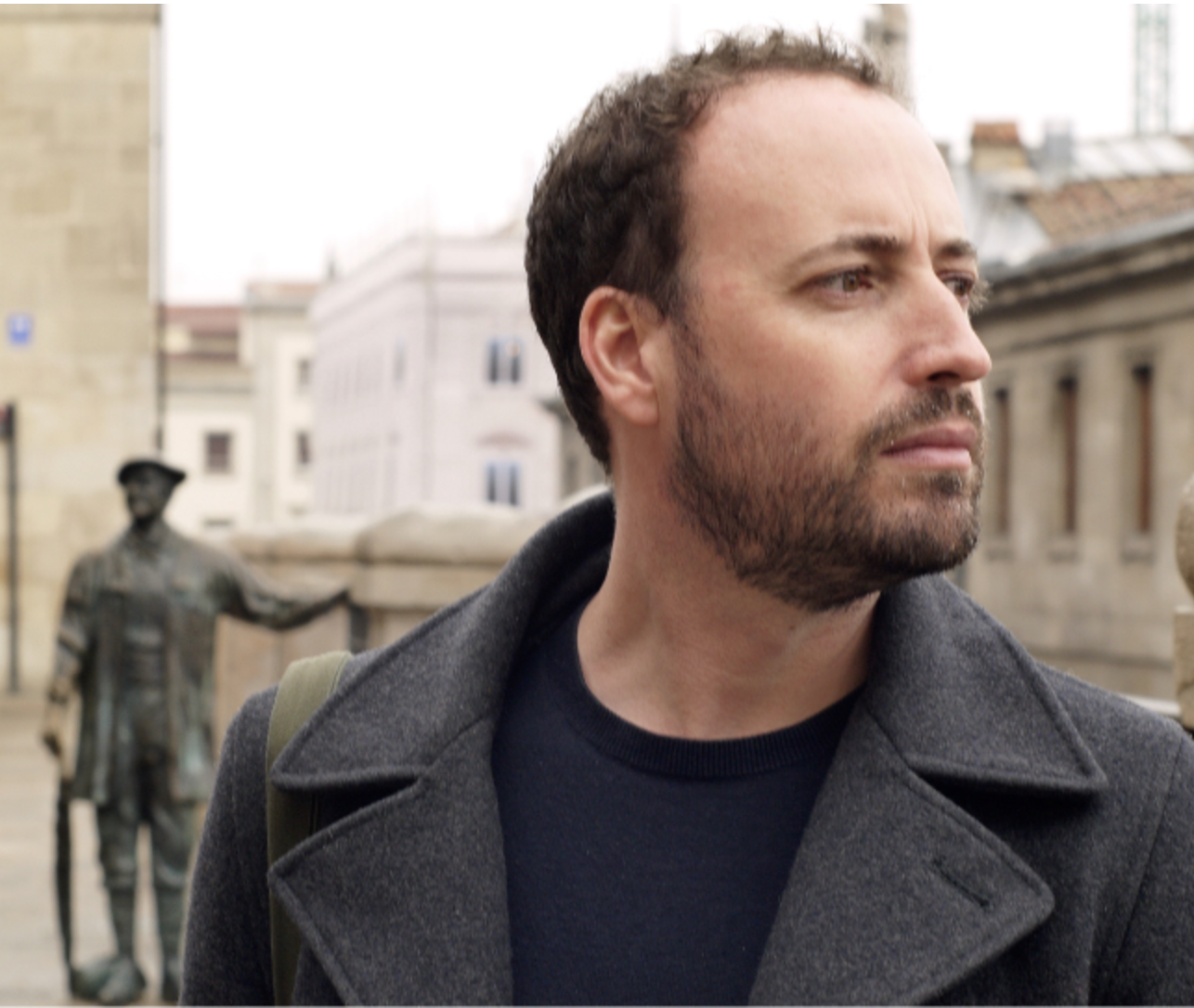 Anthony Gardner profile photo. Anthony is looking to the right, wearing a dark grey coat with a navy roundneck t-shirt. In the background there is a sculpture of a man with one hand resting on a wall