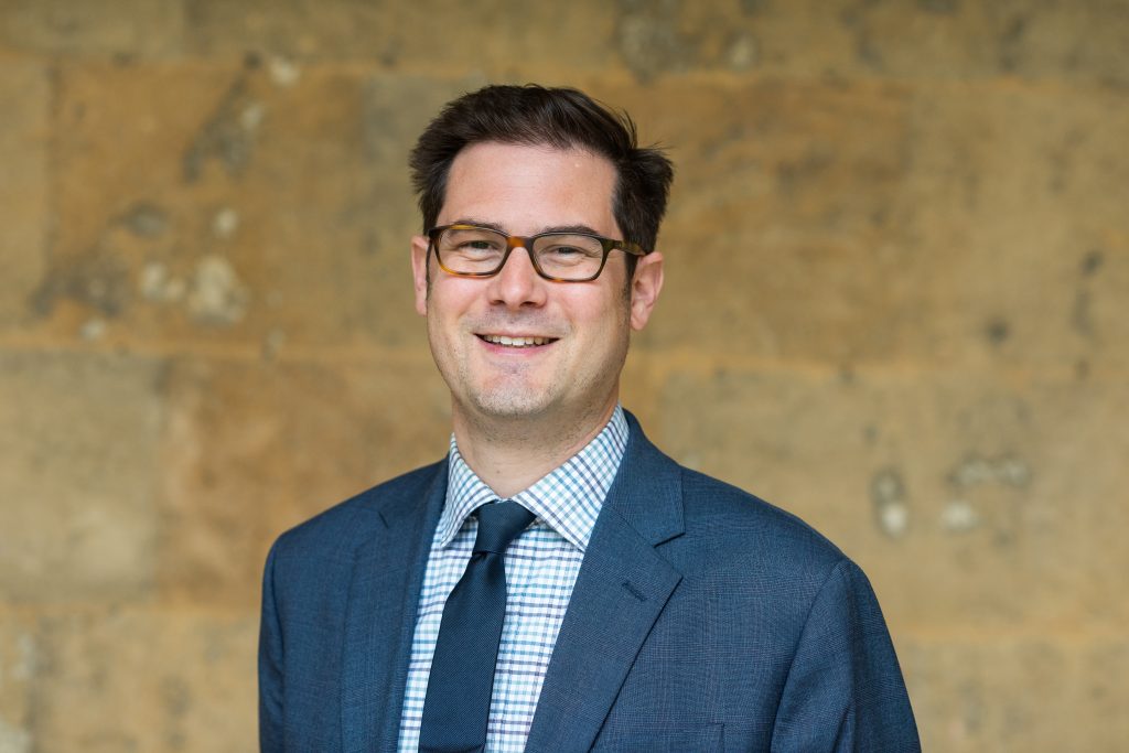 Justin Jacobs' profile photo. Justin wears a blue suit and blue tie and stands in front of the College's stone walls