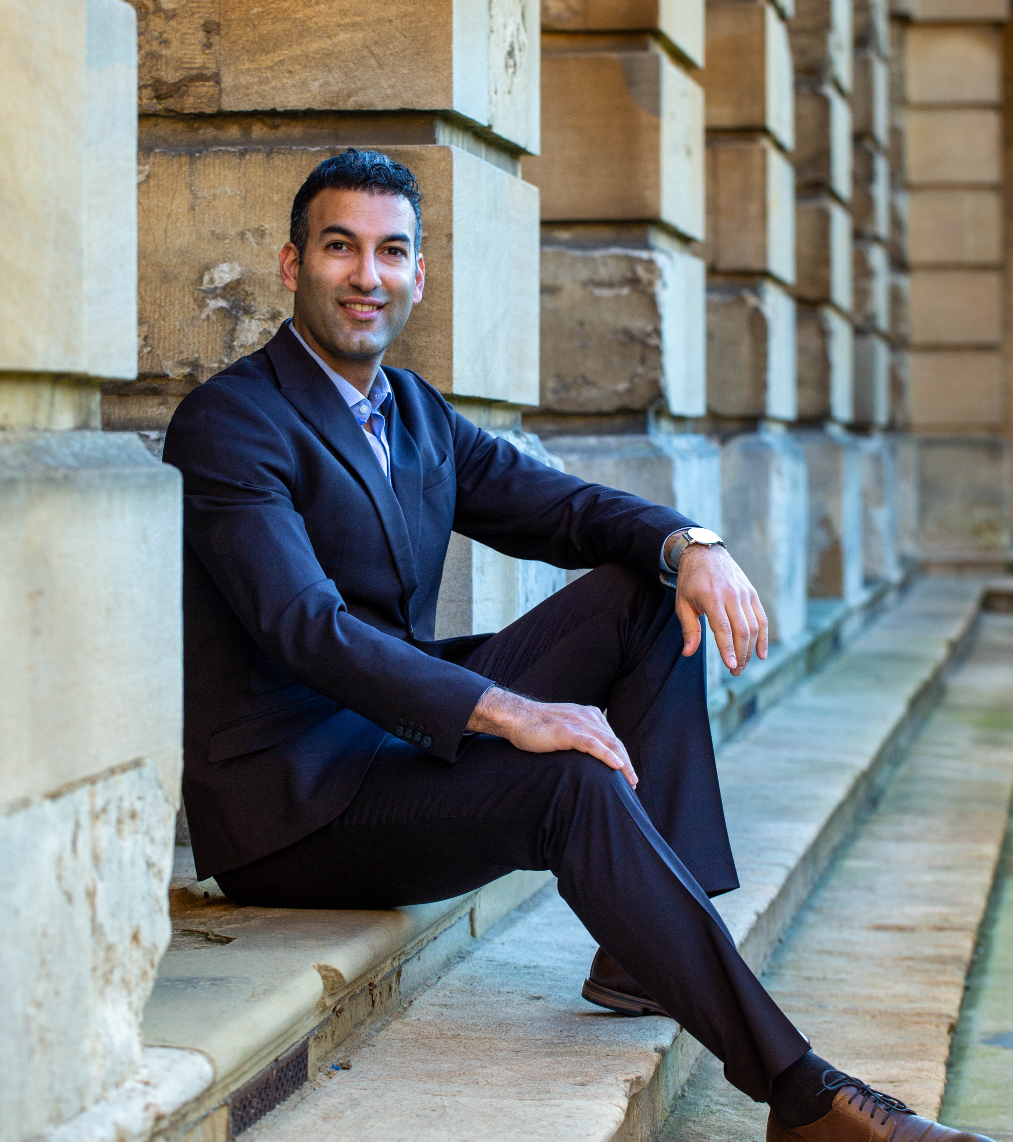 Dr Farsan Ghassim profile photo: Farsan sits on the steps of the Front Quad at Queen's College. He wears a dark suit and brown shoes