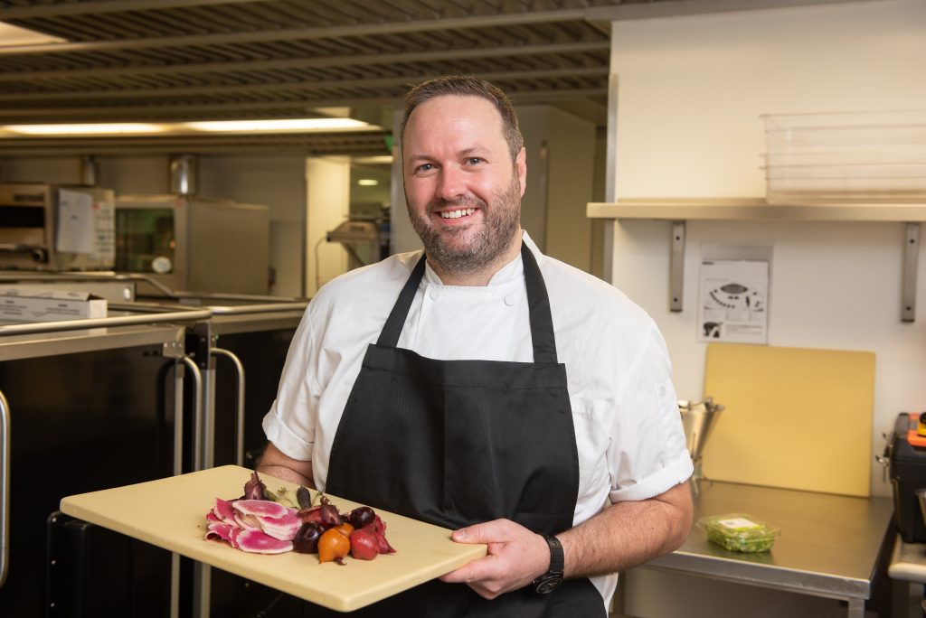 Sean pictured in the College kitchen holding a chopping board with sliced vegetables and smiling to camera