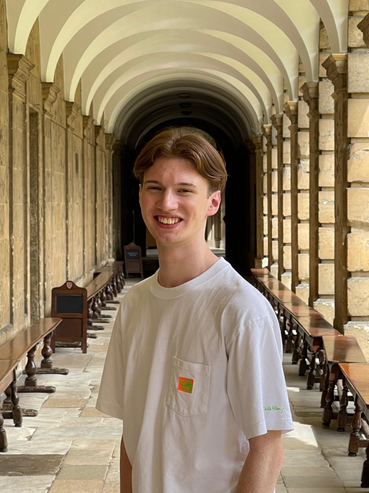 Samuel Wood, a young man with brown hair in a middle part, stands centre frame in the cloisters of The Queen’s College, Oxford. He is visible from the torso up, wearing a loose white t-shirt and smiling at the camera. Sunlight is filtering through the sandy-coloured columns from the right-hand side. The ceiling is arched and painted white, and the stone cloisters disappear into dark shadows behind Samuel. Polished dark-brown wooden benches line either side.