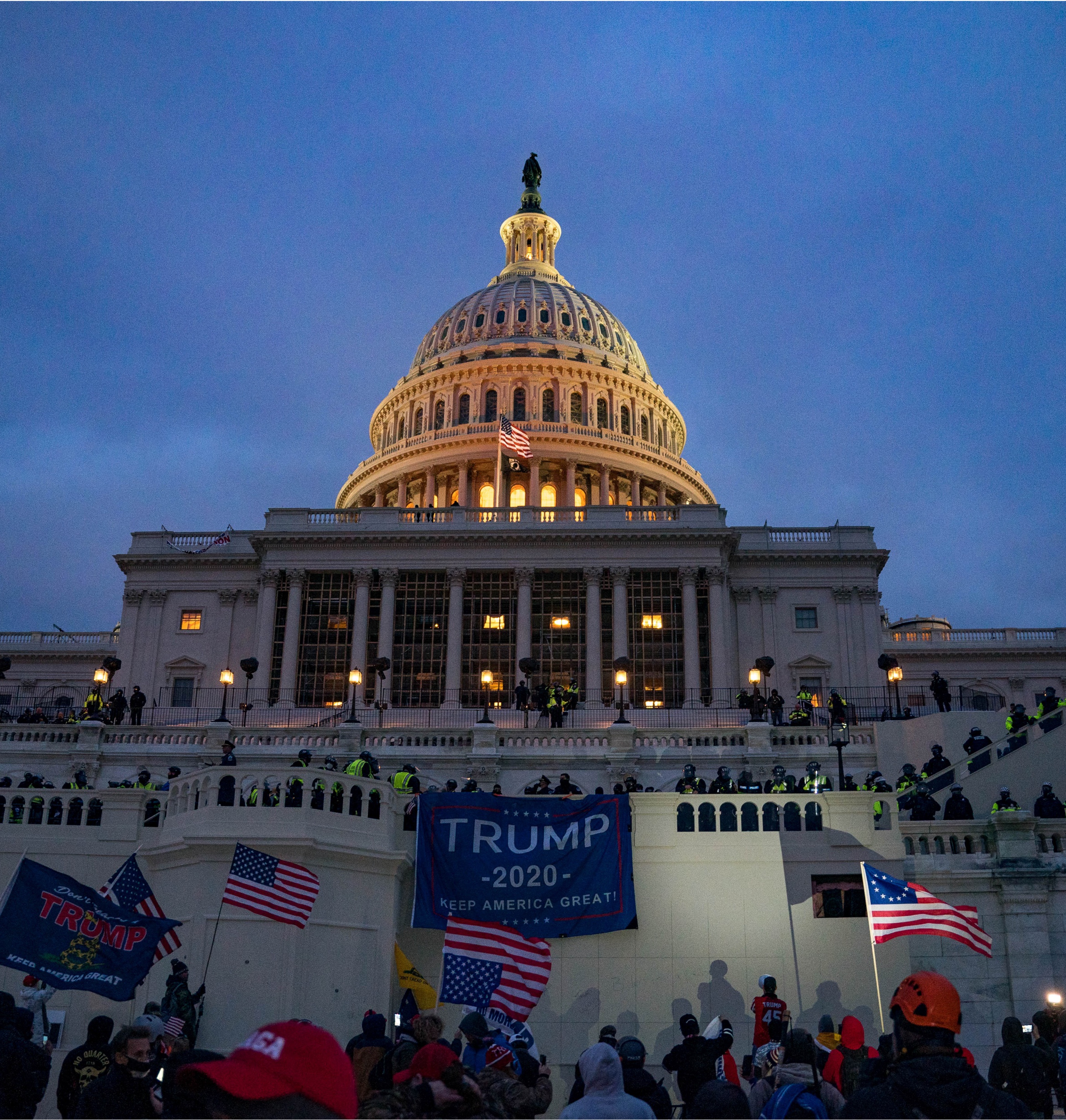 the US Capitol building with protestors waving American flags and a 'Trump 2020' banner. It is twilight and there's a heavy police presence on the upper balconies and stairs.