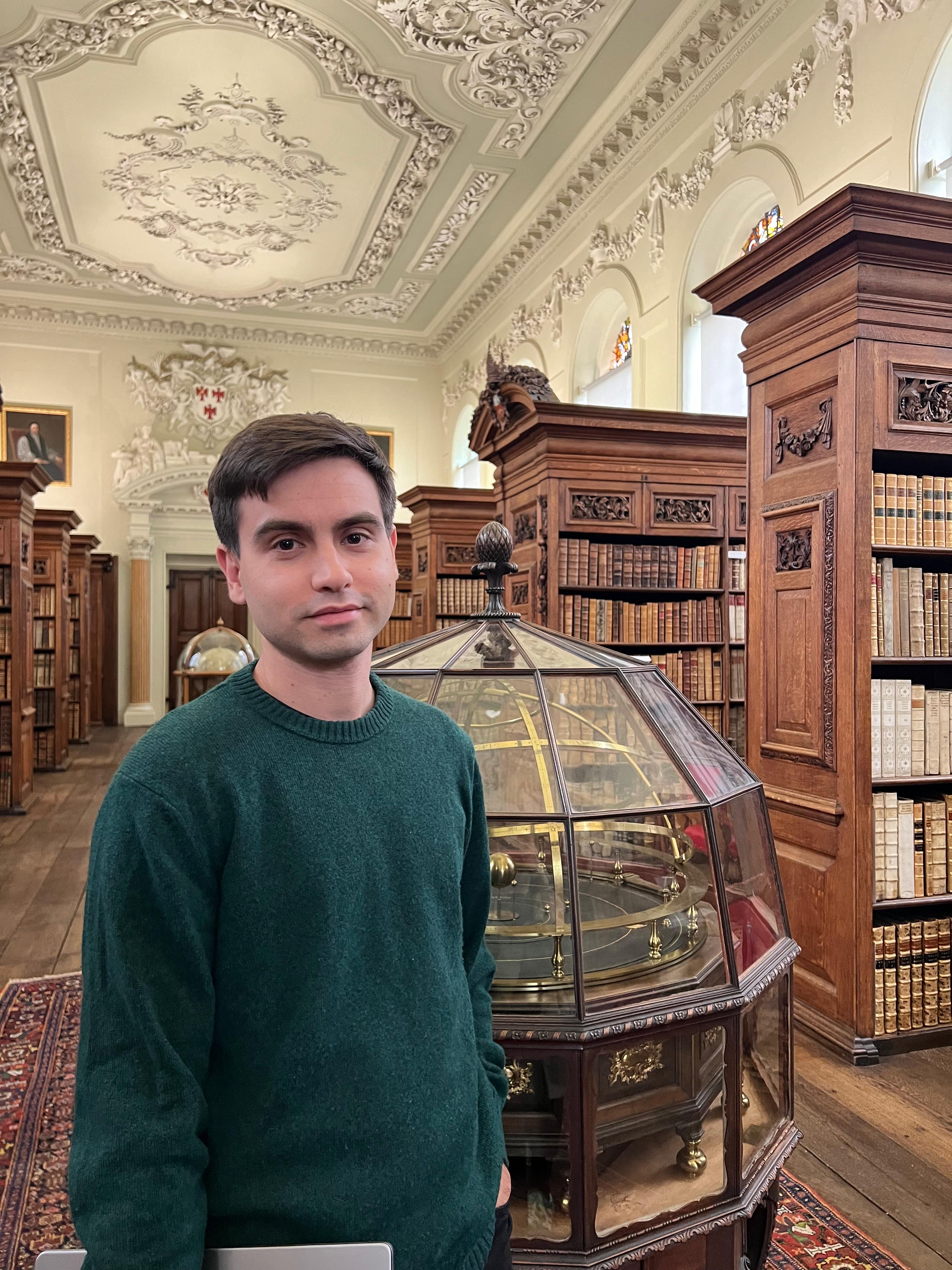 Josu Aurrekoetxea standing in the 17th century Upper Library with bookshelves and an orrery behind him. He wears a dark green jumper.