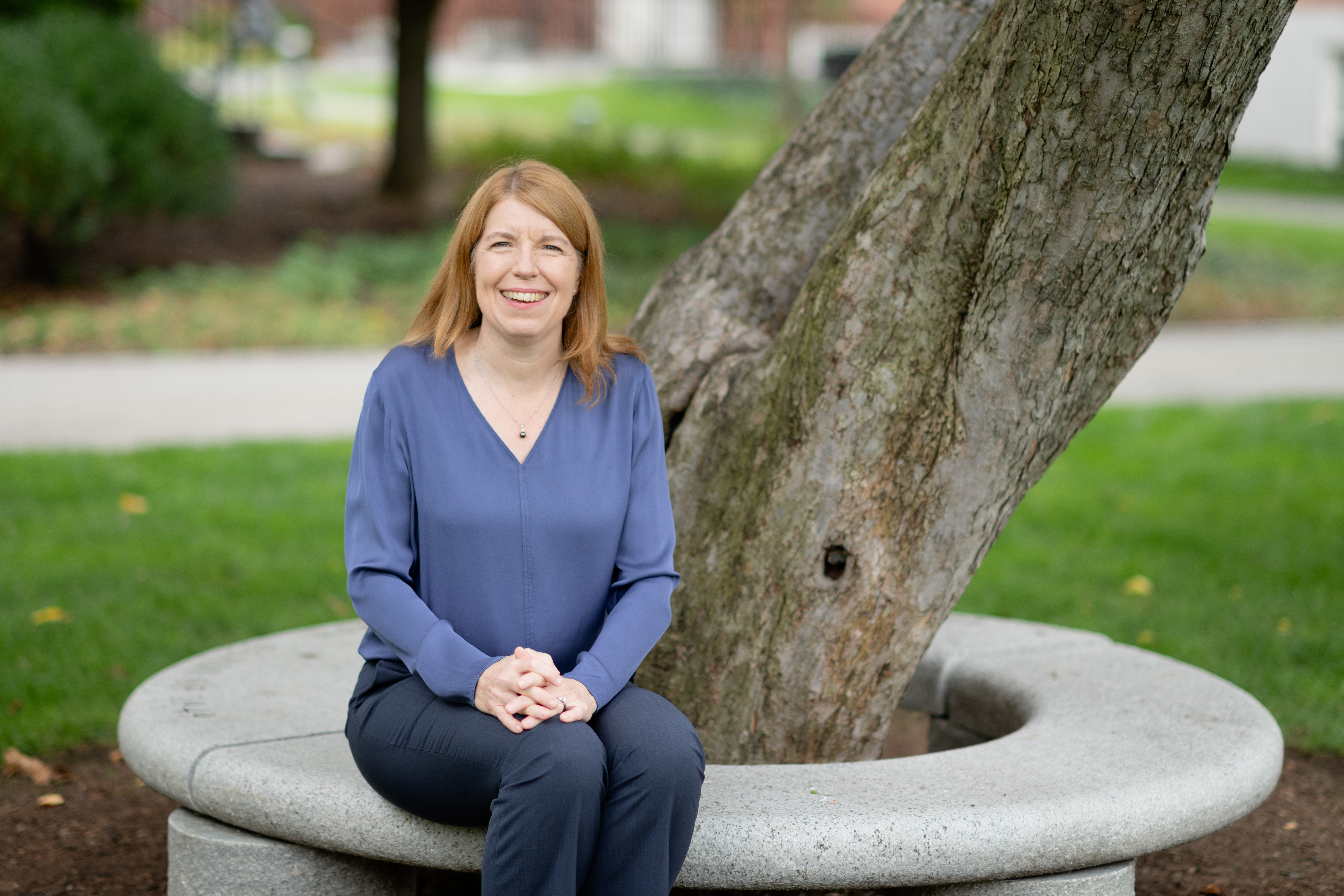 Professor Christina Davis sitting on a circular stone bench that goes around the trunk of a large tree. She is wearing a blue v-neck jumper and dark trousers.