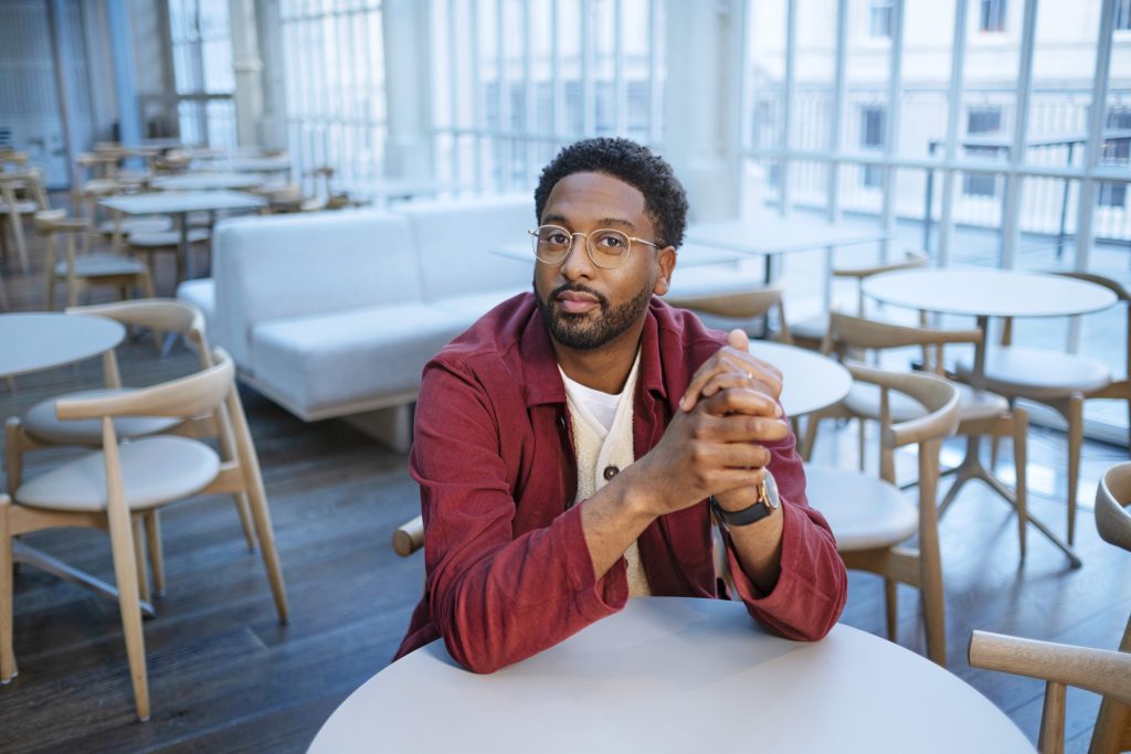 Peter Brathwaite profile image. Peter is sitting at a white circular table with his hands clasped and elbows on the table. He is wearing a red shirt. The room behind him is full of tables and Scandi-style chairs. The windows let in lots of cool blue light.