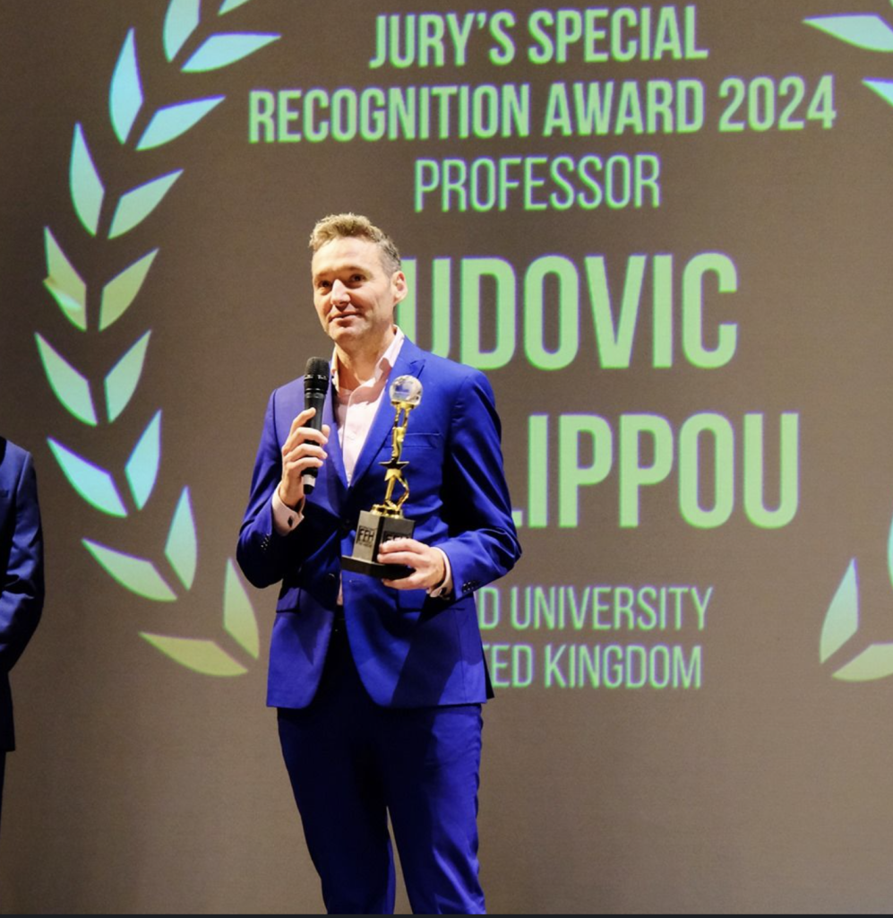 Prof Phalippou pictured in a right blue suit at the film awards holding a microphone and an award. He is standing in front of a screen which has his name and the award title in large green letters