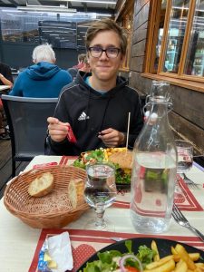 Sam sitting at a table for two opening a packet of ketchup. There's plate of food in front of him with a burger, chips and salad on it.