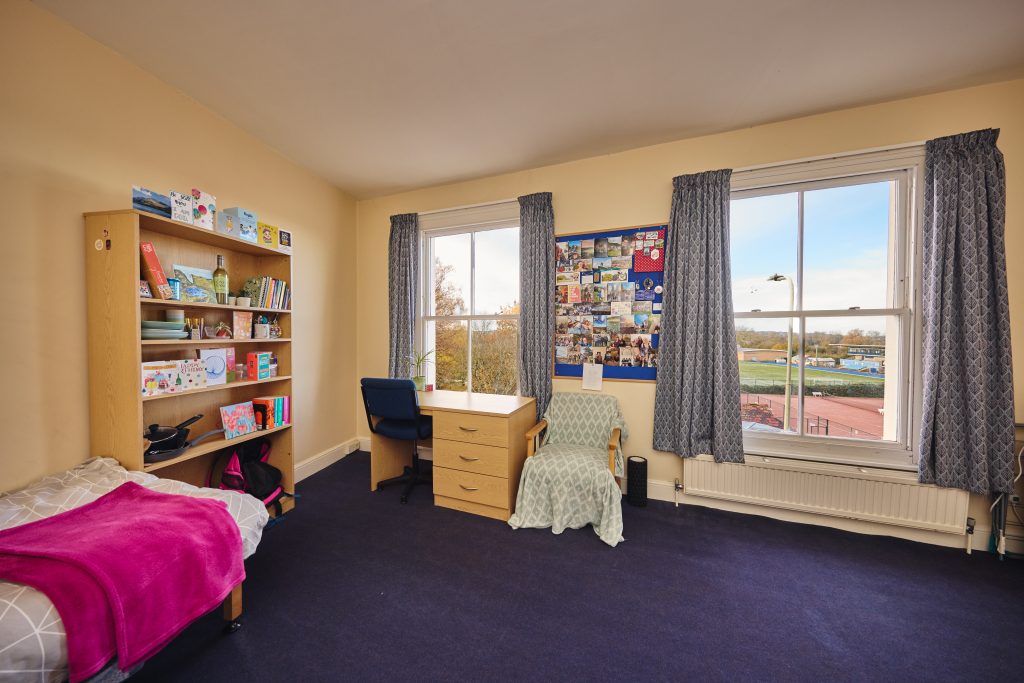 Cardo bedroom with bed, tall bookshelf, desk, chair and drawers, a comfy chair and two large window showing the running track across the road