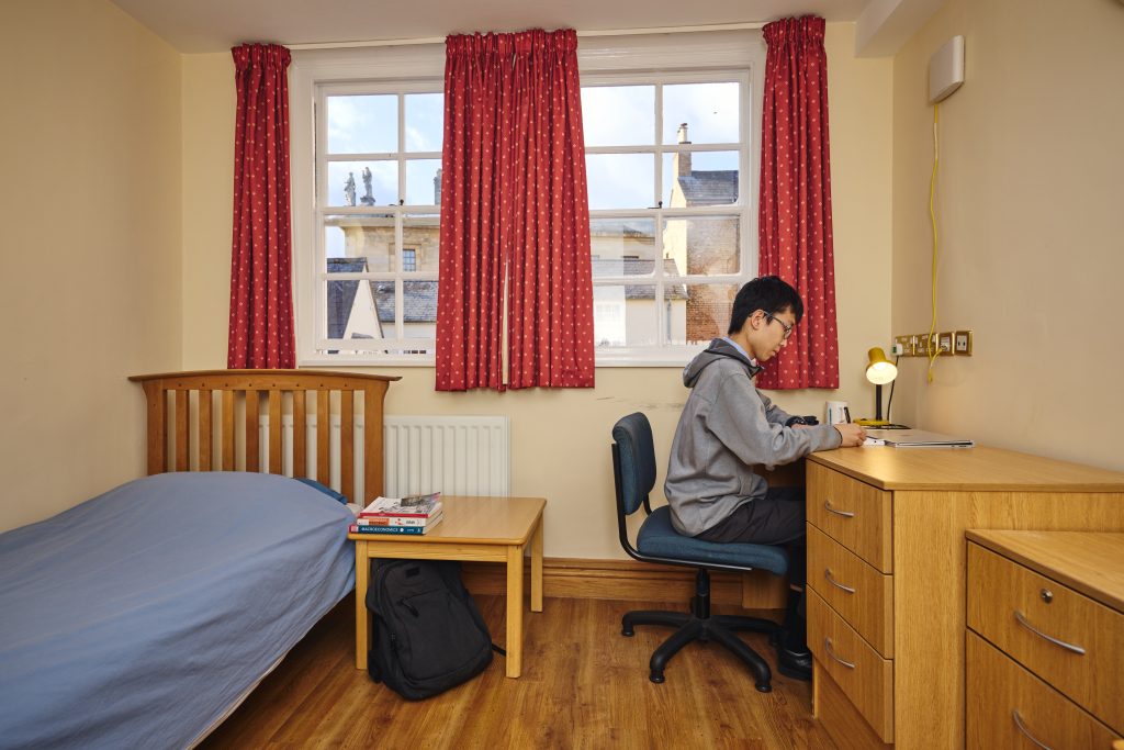 student bedroom with bed, small table, desk, drawers and two windows. A student sits at his desk writing. Through the window you can see the Queen's College main building