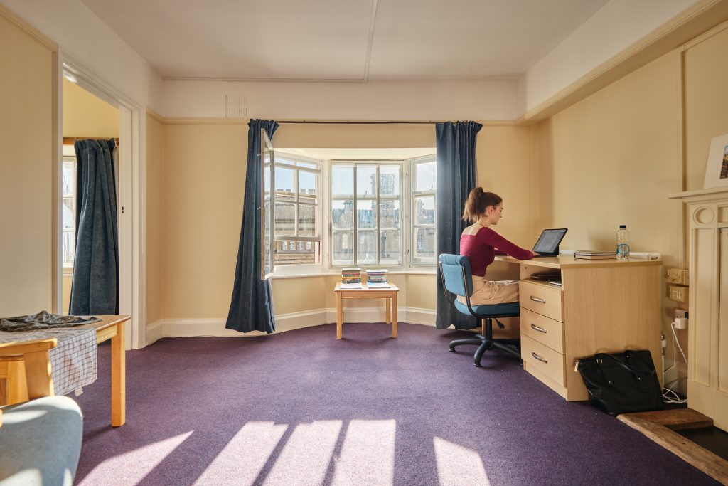 A student bedroom with a purple carpet, large bay window and desk to the right. A student sits at her laptop working. Sunlight pours through the window which has a small coffee table with books piled up.