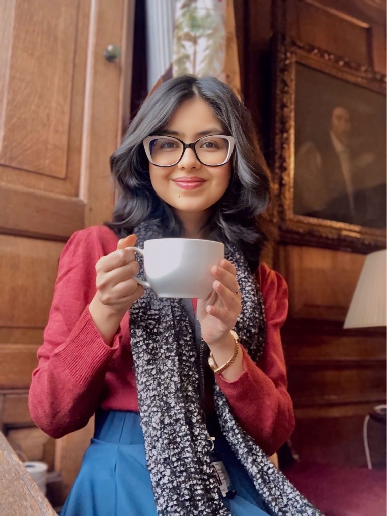 Profile photo for Jennice Singh: she sits with a cup of coffee with wooden panelling and portraits behind her