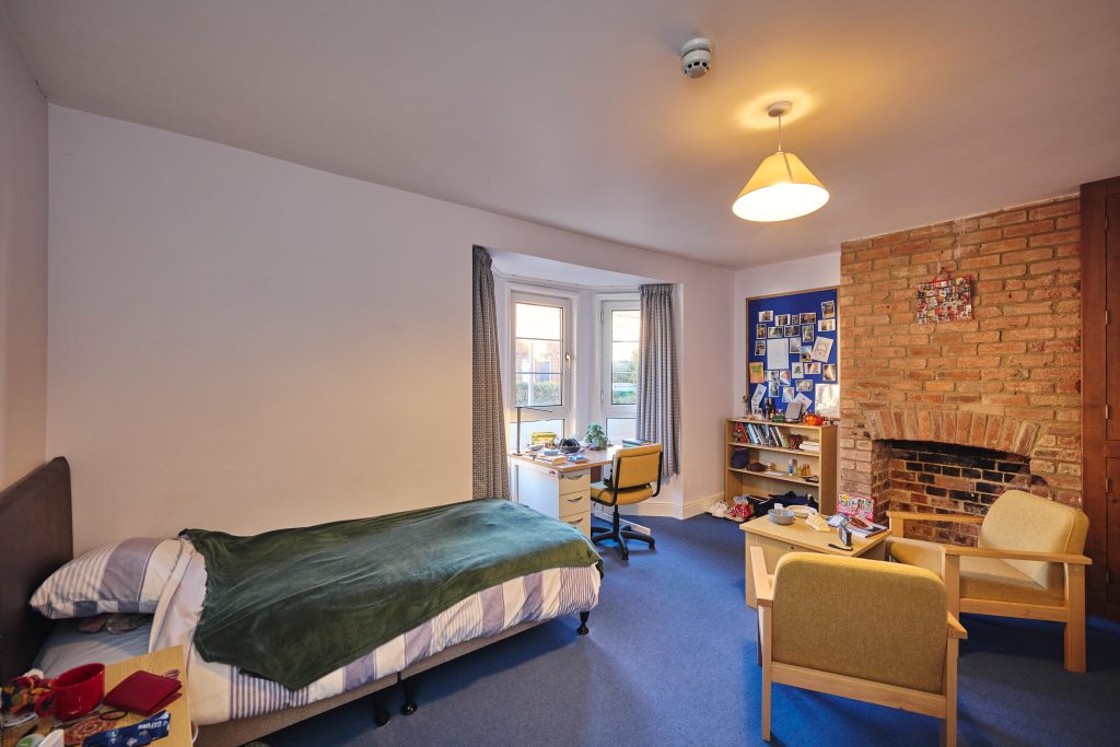 a ground floor student bedroom showing a bed, two chairs round a coffee table, a brick fireplace, a large window and a desk area with pinboard behind.