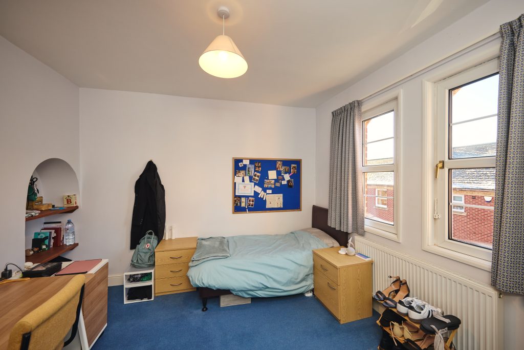 a first floor student bedroom showing a bed, small bedside table, mini chest of drawers, a small alcove with shelves, a desk and chair and a shoe rack. The room has two windows and a pinboard on the wall.