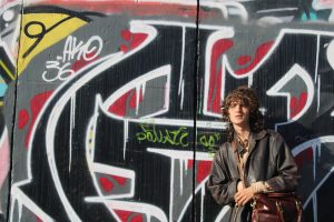 Kit's profile photo: he stands in front of a wall covered in graffiti