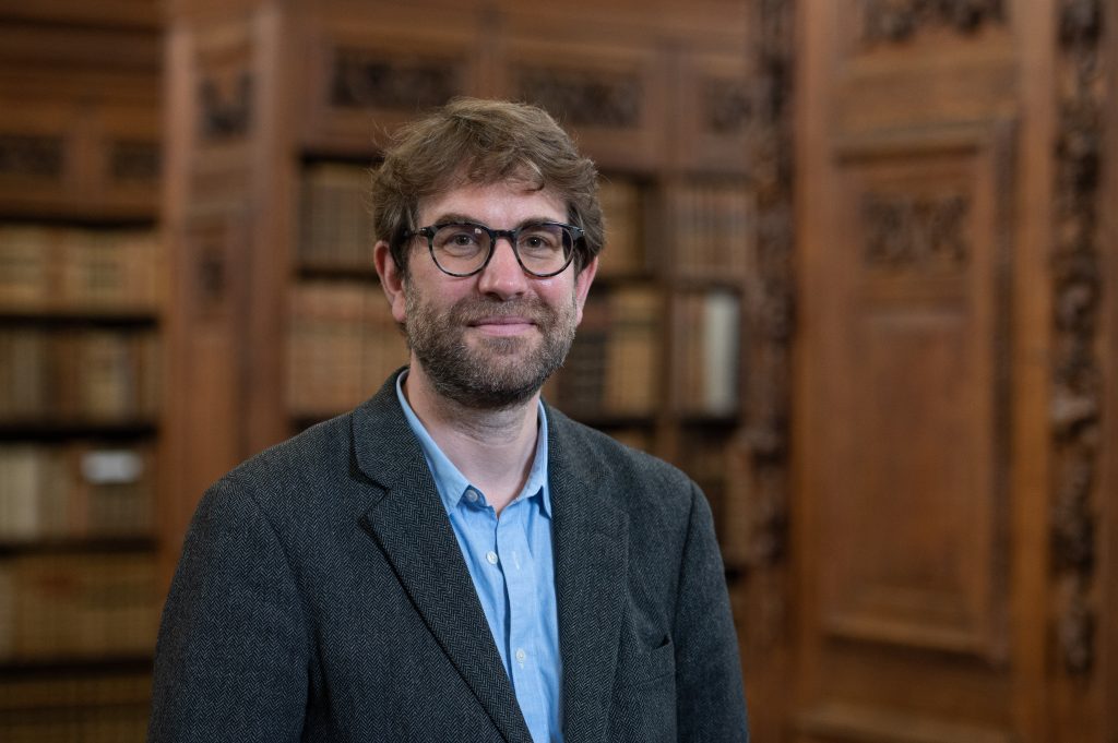 Matt Shaw pictured in the Upper Library wearing a blue shirt and jacket standing in front of wooden bookcases