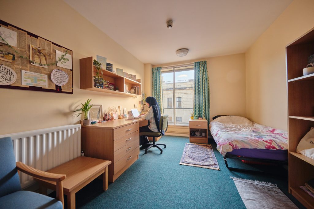 Student bedroom at St Aldate's House. There's a bed, tall bookshelf, chest of drawers, comfy chair, low table, desk and chair. A student sits at the desk typing. There's a large window at the end of the room.