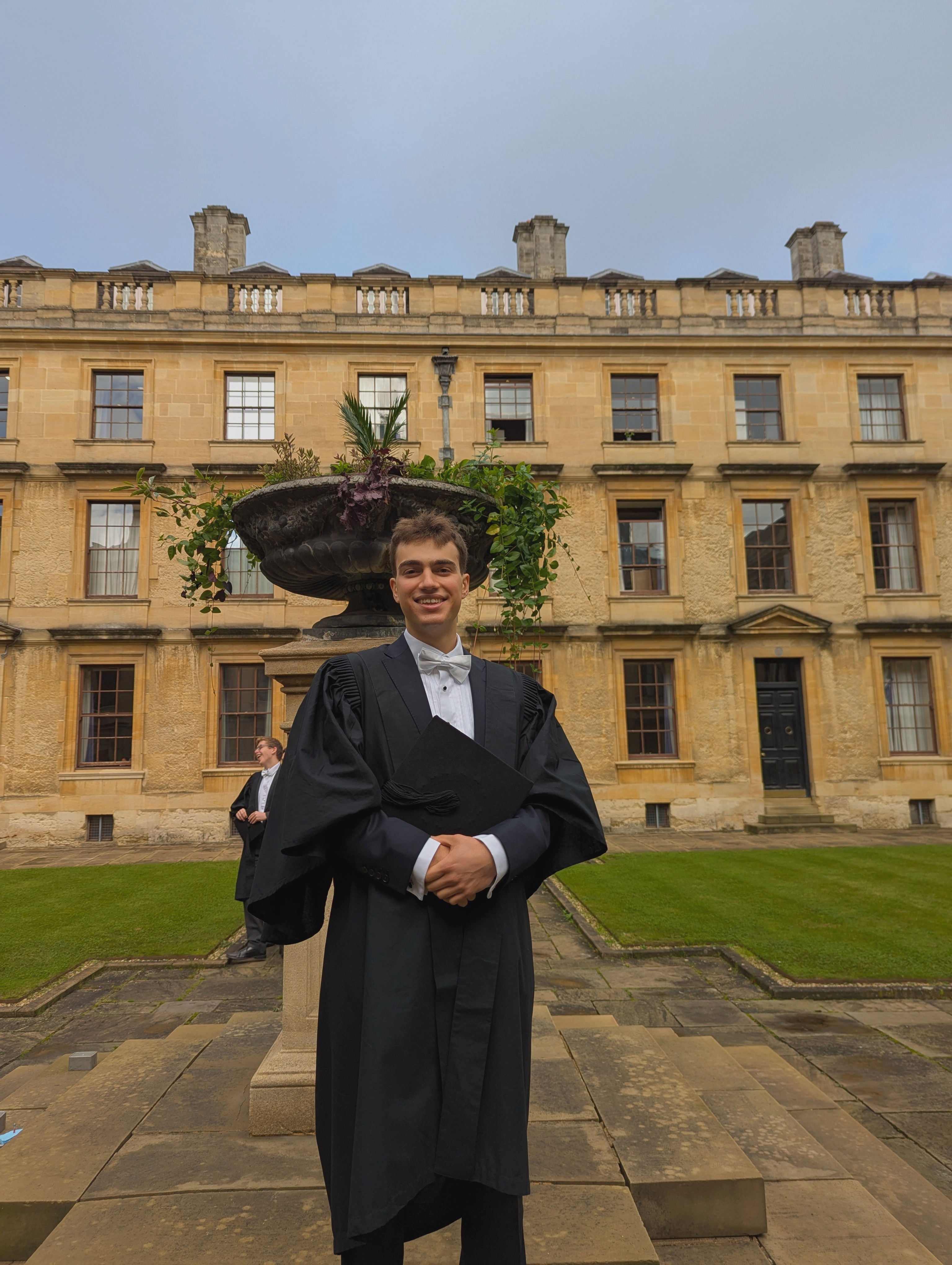 Tom's profile photo taken in Back Quad. He wears a black scholar's gown and holds a mortarboard