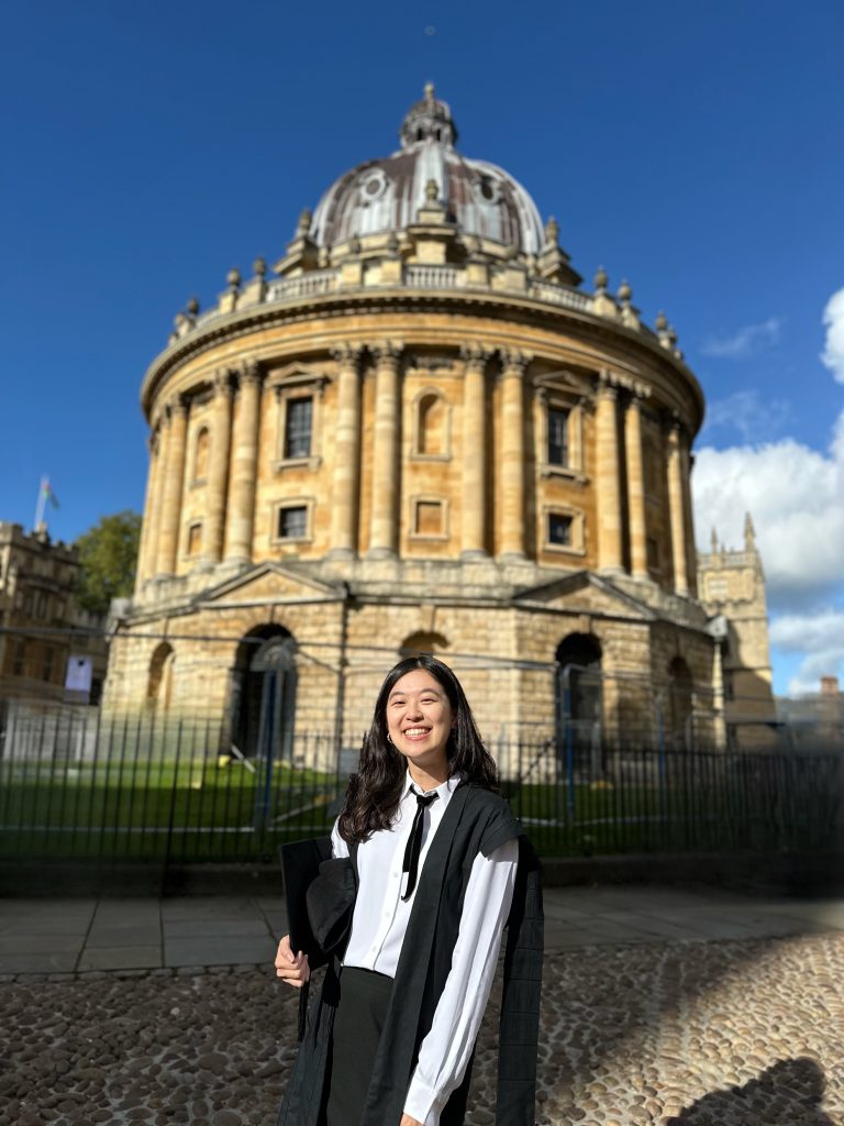 Profile photo of Vivi wearing her subfusc (black gown, white shirt and black ribbon tie) in front of the Radcliffe Camera library building