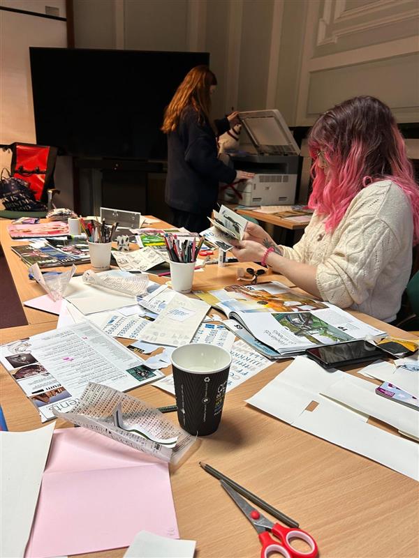 Library staff at the zine-making workshop. The desk is covered with clippings, scissors, and pots of pens