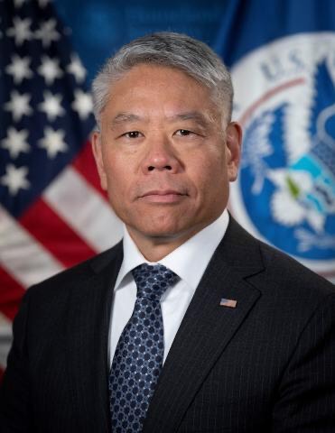 John Tien profile photo. John wears a dark suit and blue tie and stands in front of the US flag.